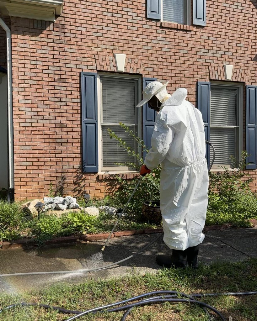Vulcan technician in white protective suit pressure washing a sidewalk beside a brick home.